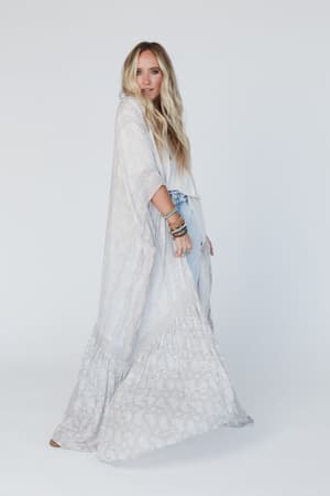 A woman with long blonde hair wears the Paisley Tapestry Free Flow Duster Kimono in gray over a white top and blue jeans, accessorized with bracelets, standing against a plain white background.