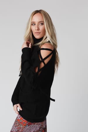 A woman with long blonde hair wears the Roxy Criss Cross Tie Shoulder Sweater in black, featuring sleeve cutouts and laces, paired with a colorful patterned skirt against a plain light background.