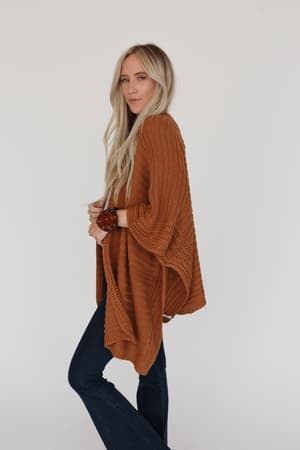 A woman with long blonde hair models the So Deserving Knit Ruana in camel, styled open over dark jeans. She stands sideways against a white background, looking at the camera with a relaxed vibe—ideal for effortless boho outfits.
