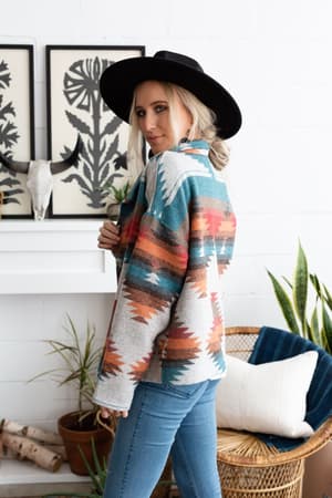 A woman in the Anika Mixed Pattern Jacket - Teal and a black hat stands indoors, looking over her shoulder amid plants, a wicker chair with a pillow, and Southwestern decor on a white wall.