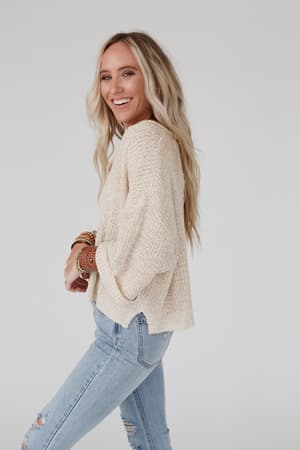 A woman with long blonde hair wears the Just Right Slouchy Sweater in oatmeal, paired with ripped light blue jeans and beaded bracelets, smiling while standing sideways against a plain white background.