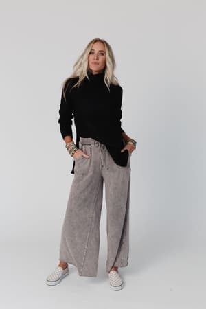 A woman with long blonde hair stands against a plain background, wearing the Danny Sweater in black, loose gray wide-leg pants, checkered slip-on shoes, and colorful bracelets. Her hands are in her pockets as she looks confidently ahead.