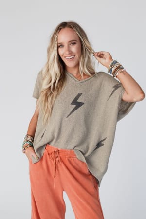 A smiling woman with long blonde hair wears the Struck By You Dolman Sleeve Sweater Top in mocha, styled with orange drawstring pants and colorful beaded bracelets. She stands against a plain light background.
