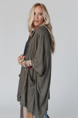A woman with long blonde hair poses in The Nest Boho Essential Hooded Ribbed Sweater in Olive over a black top, accessorized with colorful bracelets, standing against a plain light background.