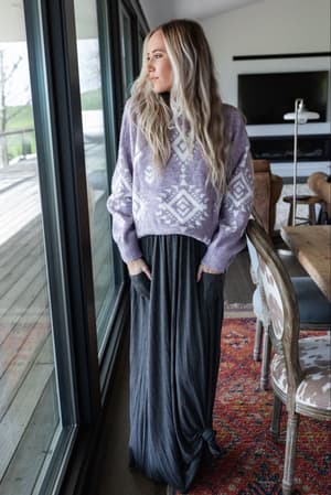 A woman with long blond hair, in a purple patterned sweater and The Perfect Pocketed Maxi Skirt - Charcoal, stands by large windows in a modern room with a rug, dining table, and chairs, gazing outside.