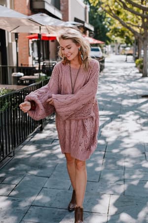 A woman walks on a sunlit sidewalk wearing the Golden Hour Bubble Sleeve Sweater in mauve styled as a dress with brown ankle boots, smiling with trees and outdoor seating in the background.