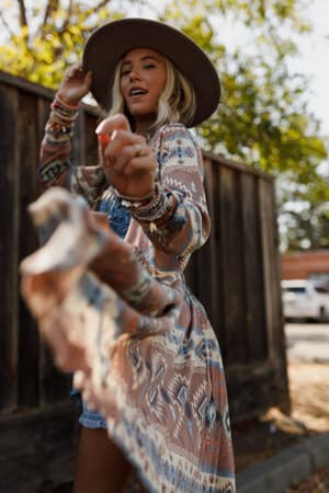 Multi / A woman wearing a Santa Fe Rose Duster Cardigan with a boho print, paired with a blue dress and tan hat, standing against a wooden fence.