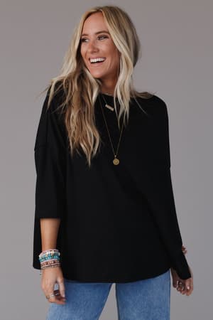 A smiling woman with long blonde hair wears The Nest Cozy Cool Oversized Tee in black, styled with light blue jeans, layered necklaces, and colorful bracelets, standing against a plain grey background.