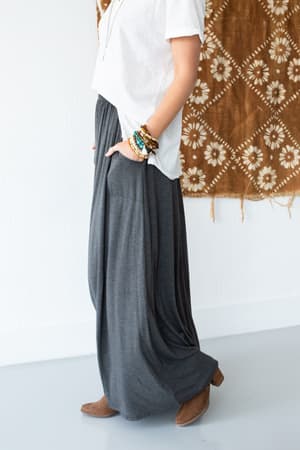 A person wearing a white shirt, The Perfect Pocketed Maxi Skirt - Charcoal, brown ankle boots, and colorful bracelets stands in front of a patterned brown textile hanging on a white wall.