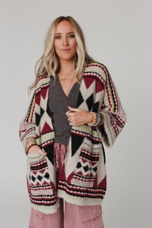 A woman with long blonde hair wears the Keep It Up Oversized Cardigan - Berry, featuring geometric designs in red, black, and cream over a dark top and pink pants. She accessorizes with bracelets and stands against a light gray background.
