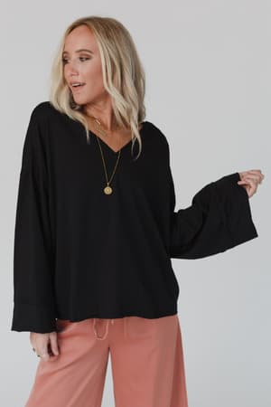 A woman with long blonde hair wears The Nest Love It V Neckline Sweater Top in black and pink pants. She looks to her left, accessorized with a gold pendant necklace, against a plain light background.