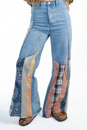 A person wearing Magnolia Patchwork Jeans - Washed Denim with colorful panels and frayed hems, paired with brown sandals. The image is cropped at the waist, showing only the lower half.