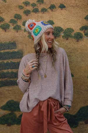 A woman with long blonde braids smiles, wearing a colorful crocheted beanie, rust-colored pants, and The Nest Fly With Me Batwing Sleeve Knit Sweater in Sand. She stands with one hand in her pocket before a mural of green trees on a tan wall.