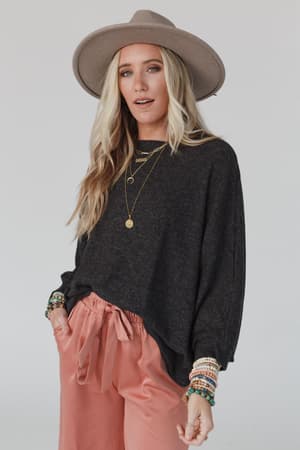 A woman in The Nest Fly With Me Batwing Sleeve Knit Sweater - Charcoal, a wide-brimmed hat, pink satin pants, layered necklaces, and beaded bracelets stands against a plain light background.