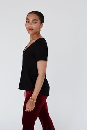 A woman with tied-back hair wears the Scoop Neck Bralette Tee in black and red velvet pants, standing against a plain light background and looking over her shoulder with a slight smile.