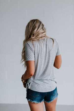 A person with long, wavy blonde hair stands with their back to the camera, wearing the Scoop Neck Bralette Tee - Gray and blue frayed denim shorts against a plain light-colored background.