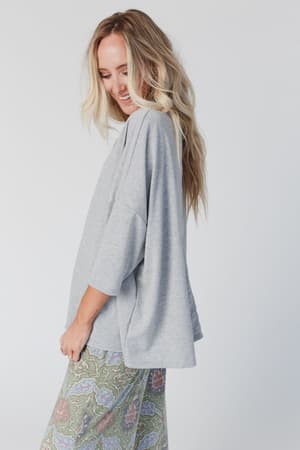A smiling woman with long, wavy blonde hair looks down, wearing The Nest Cozy Cool Oversized Tee in Heather Gray and patterned pastel pants. She stands against a plain light background, creating a relaxed, boho vibe.