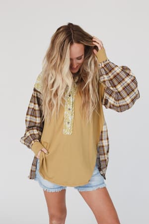 A woman with long blonde hair wears the Better Together Mixed Print Top - Mustard with light denim shorts, standing against a white background, looking down and smiling with one hand in her hair.