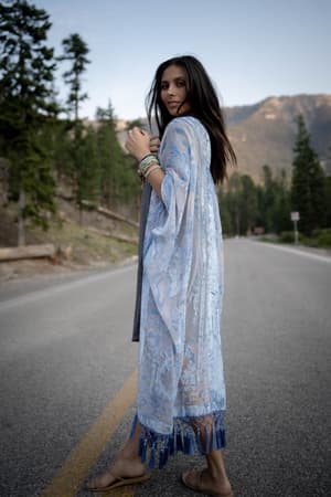 A woman in the Crushin' On You Velvet Kimono - Light Blue stands barefoot on an empty road, surrounded by tall trees and distant mountains under a clear sky.