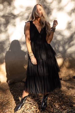 A woman in the Boho Ballerina Tiered Tulle Dress - Black and boots stands on leaf-covered ground in sunlight. With long, wavy blonde hair and stacked bracelets, she embodies effortless boho-chic style.