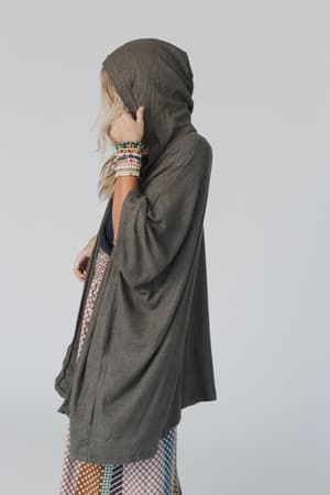 A woman with long blonde hair wears The Nest Boho Essential Hooded Ribbed Sweater in olive as a loose cape, along with colorful beaded bracelets and a patterned skirt. She stands sideways against a light gray background, her hood and hand partly covering her face.