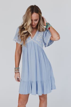 A woman with long blonde hair wears the Magdaline Mini Dress - Blue, a light blue short-sleeved embroidered mini dress with a ruffle hem. She looks down, touching her hair and wearing colorful bracelets against a plain white background.