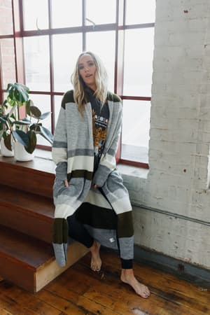 A barefoot woman with long blonde hair sits on wooden stairs by a large window, wearing The Nest So Cozy Oversized Striped Knit Duster in Olive Multi over a graphic tee and black pants. A potted plant is nearby.