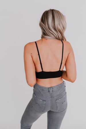 A woman with long, wavy blonde hair stands facing away, wearing the Audra Waistband Loop Lace Bralette in black paired with high-waisted gray jeans against a plain white background.