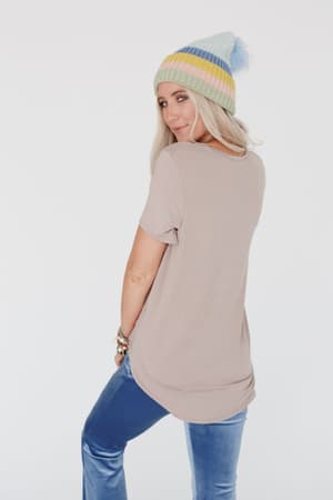 A woman with long blonde hair wears a pastel-striped beanie, blue velvet pants, and the Scoop Neck Bralette Tee in Khaki. She stands sideways, looking over her shoulder against a plain white background.