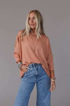 A woman with long blonde hair wears The Nest Cozy Cool Oversized Tee in coral, paired with light blue high-waisted jeans and layered jewelry, standing against a plain gray background with one hand in her pocket.