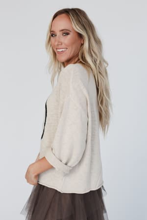 Woman with long blonde hair wears the Made You Smile Sweater in ivory, paired with a dark tulle skirt. She smiles and looks over her shoulder against a plain light background.