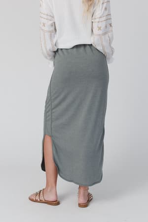 Only the lower half is visible as a person stands facing away, wearing a white patterned top and The Nest So Comfy Drawstring Maxi Skirt in Light Olive, featuring an elastic waistband, side slit, and paired with studded sandals.