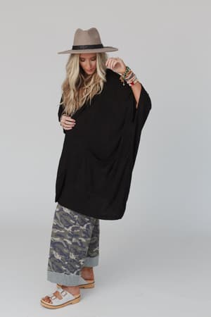 Wearing The Nest Boho Essential Hooded Ribbed Sweater in black, a woman in a wide-brimmed hat, camo wide-leg pants, white sandals, and colorful bracelets stands against a plain background, looking down and touching her hat.