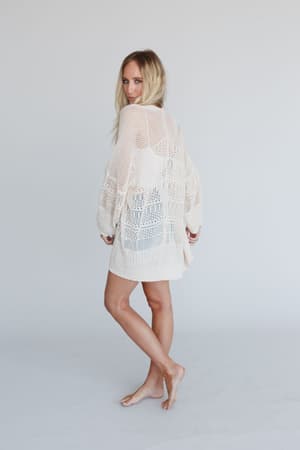 A woman with long blonde hair stands barefoot against a light background, wearing the Brenden Knit Netted Cardigan in Ivory. The loose-fitting cardigan reaches mid-thigh as she looks over her shoulder and smiles at the camera.