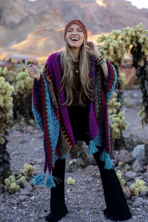 A woman smiles outdoors in a desert landscape, wearing the Desert Dreams Crochet Ruana - Magenta from her boho closet, black pants, bracelets, and a reddish headband. Cacti and rocky terrain appear in the background.