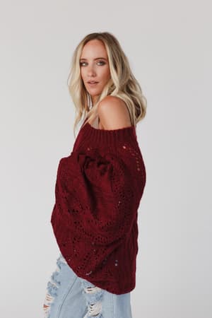 A woman with long blonde hair wears the Golden Hour Bubble Sleeve Sweater Dress in burgundy, standing against a white background and looking at the camera with a relaxed expression.