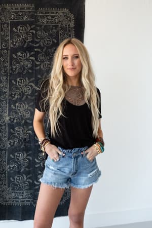 A woman with long blonde hair wears the Scoop Neck Bralette Tee - Black and ripped denim shorts, standing in front of a decorative black and white tapestry with her hands in her pockets and multiple bracelets on her wrists.