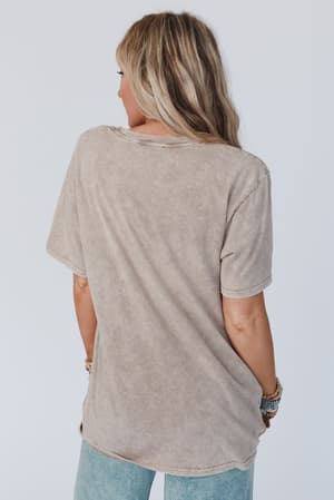 A woman with blonde hair faces away, showing off the back of her mocha Fly Free Graphic Tee and light blue jeans. Several bracelets adorn her left wrist, all set against a simple light backdrop.