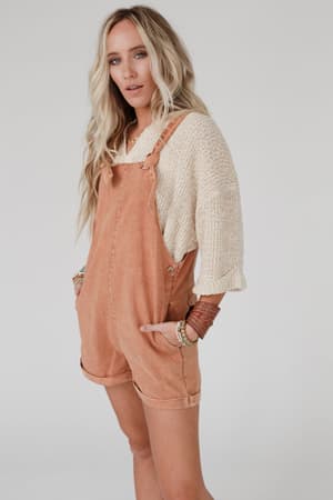 A woman with long, wavy blonde hair stands with hands in pockets, wearing the Just Right Slouchy Sweater in oatmeal under peach short overalls. She looks at the camera against a plain light background.