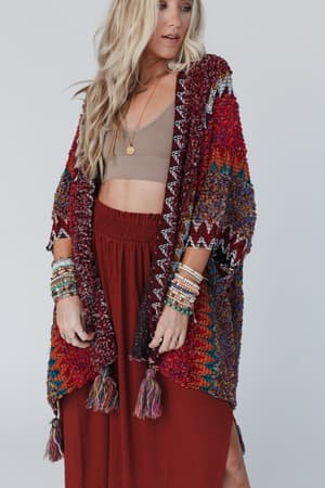 A woman models the Desert Dreams Crochet Ruana - Maroon with tassels, layered over a tan crop top and rust-red skirt. She accessorizes with beaded bracelets, capturing a boho vibe against a simple backdrop.