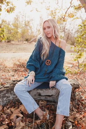 A woman with long blonde hair sits on a fallen log outdoors, surrounded by autumn leaves and bare trees, wearing the Golden Hour Bubble Sleeve Sweater Dress in teal.