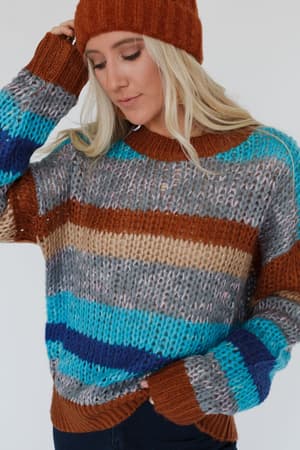 A woman stands against a plain background, looking down with one hand touching her cozy Go For It Striped Sweater - Blue and wearing a rust-colored knit beanie.