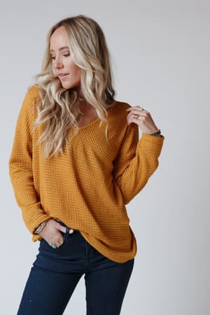A woman with long blonde hair models The Nest Flattering Open Back Thermal Knit Top in mustard with dark jeans, standing against a plain light gray background and looking down with a slight smile—cozy boho outfit inspiration.
