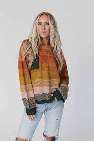 A woman with long blonde hair models the Going Places Gradient Stripe Sweater in Mustard and light ripped jeans, posing against a plain light background with one hand in her pocket.