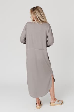 A person with long, blonde hair faces away, wearing The Nest Meadow Long Sleeve Slit Maxi Dress in coco with side slits and beige sandals, set against a plain white background.
