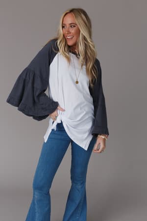 A smiling woman with long blonde hair wears the Bell Sleeve Baseball Tee in Charcoal, styled with blue flared jeans and a necklace. She poses confidently with one hand on her hip against a plain gray background, exuding effortless style.