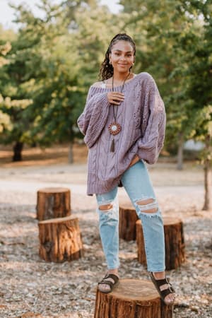 A woman smiles confidently at the camera while standing outdoors on a tree stump, wearing the Golden Hour Bubble Sleeve Sweater Dress in Lavender. Sunlight filters through the trees in the background.