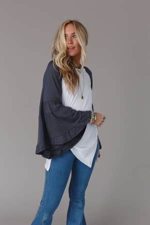 A woman with long blonde hair modeling the Bell Sleeve Baseball Tee in Charcoal stands against a plain gray background, wearing blue jeans, layered necklaces, and bracelets, and looking slightly to the side.