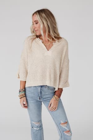 A woman with long blonde hair wears the Just Right Slouchy Sweater in oatmeal and ripped light blue jeans, accessorized with colorful bracelets, as she stands against a plain light background looking to the side.