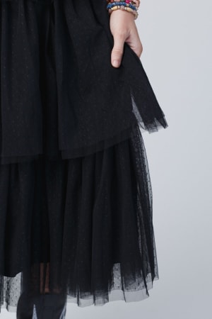 Wearing the Boho Ballerina Tiered Tulle Dress in black, a person holds the skirt with one hand. Multicolored beaded bracelets on their wrist add a boho-chic touch against a simple light gray background.